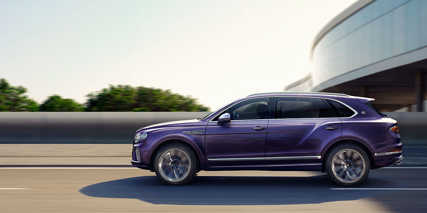 Bentley Leeds Bentley Bentayga Extended Wheelbase Mulliner luxury SUV profile in Tanzanite Purple paint, driving on an country road past trees.