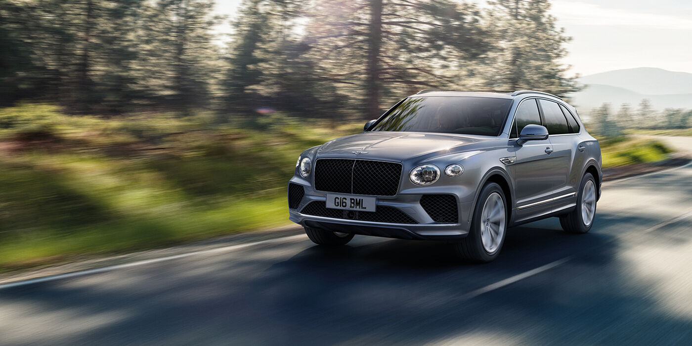 Bentley Leeds Bentley Bentayga luxury SUV front three quarter in Moonbeam paint, driving on an country road past trees.