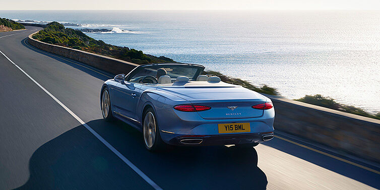 Bentley Leeds New Bentley Continental GTC Azure convertible rear view in Light Windsor blue paint with 22 inch ten spoke wheels driving dynamically on an ocean road in the sunshine
